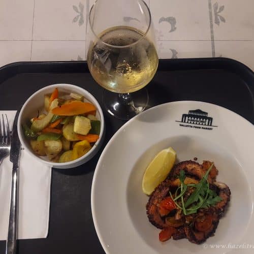 Grilled meat dish with lemon wedge, vegetable medley, and glass of white wine on a tray at Valletta Food Market, Malta. Perfect for food lovers exploring Maltese cuisine with Hazel’s Travels.