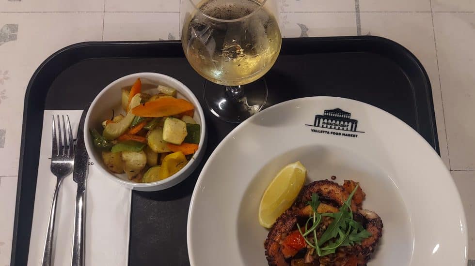 Grilled meat dish with lemon wedge, vegetable medley, and glass of white wine on a tray at Valletta Food Market, Malta. Perfect for food lovers exploring Maltese cuisine with Hazelโs Travels.