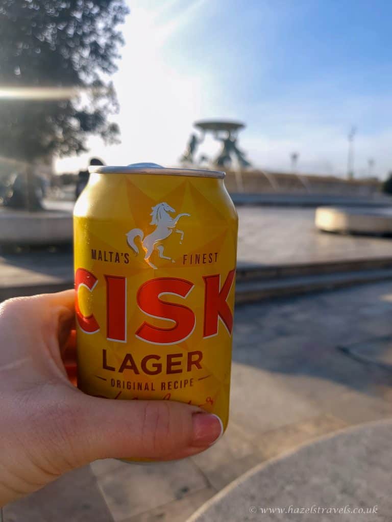 Cisk Lager can in hand with a skatepark in the background, sunny day.