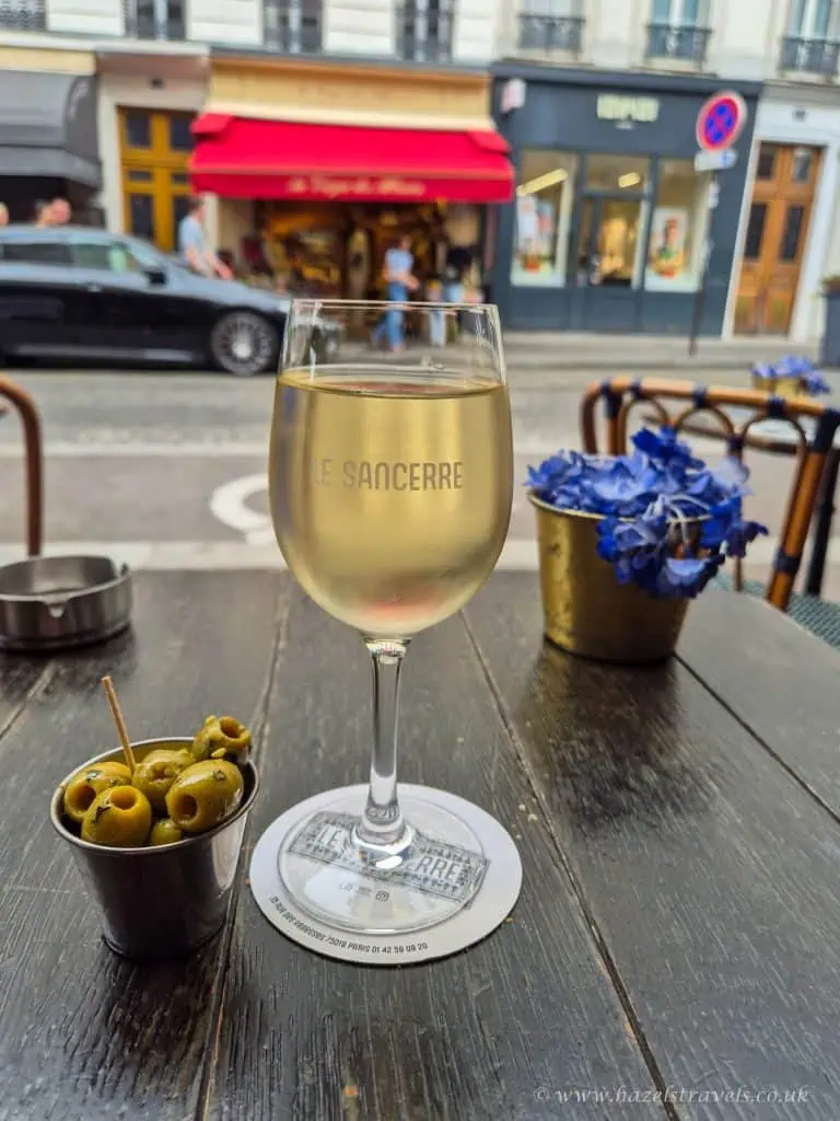 A glass of white wine and a small cup of green olives on a wooden table at an outdoor café, with a blurred street and storefronts in the background.