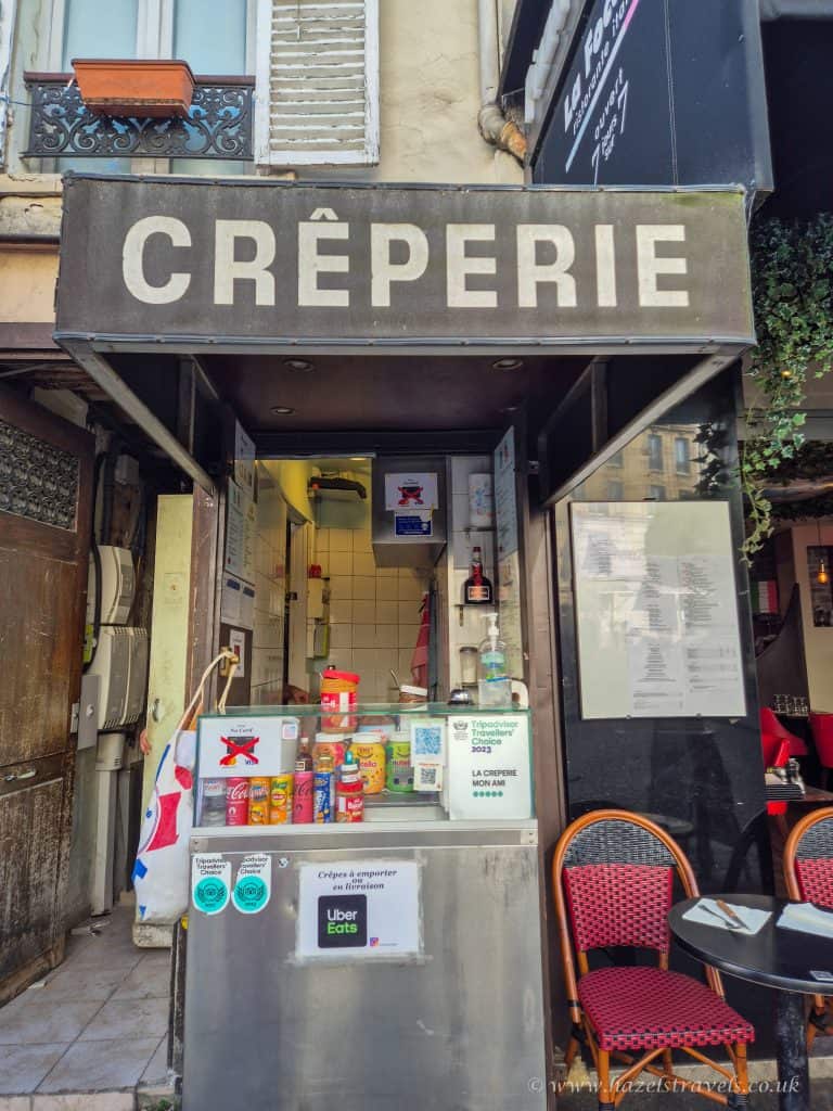 Small creperie storefront with a service counter displaying condiments and a menu, under a sign that reads "CRÊPERIE." Red chairs are visible to the right.