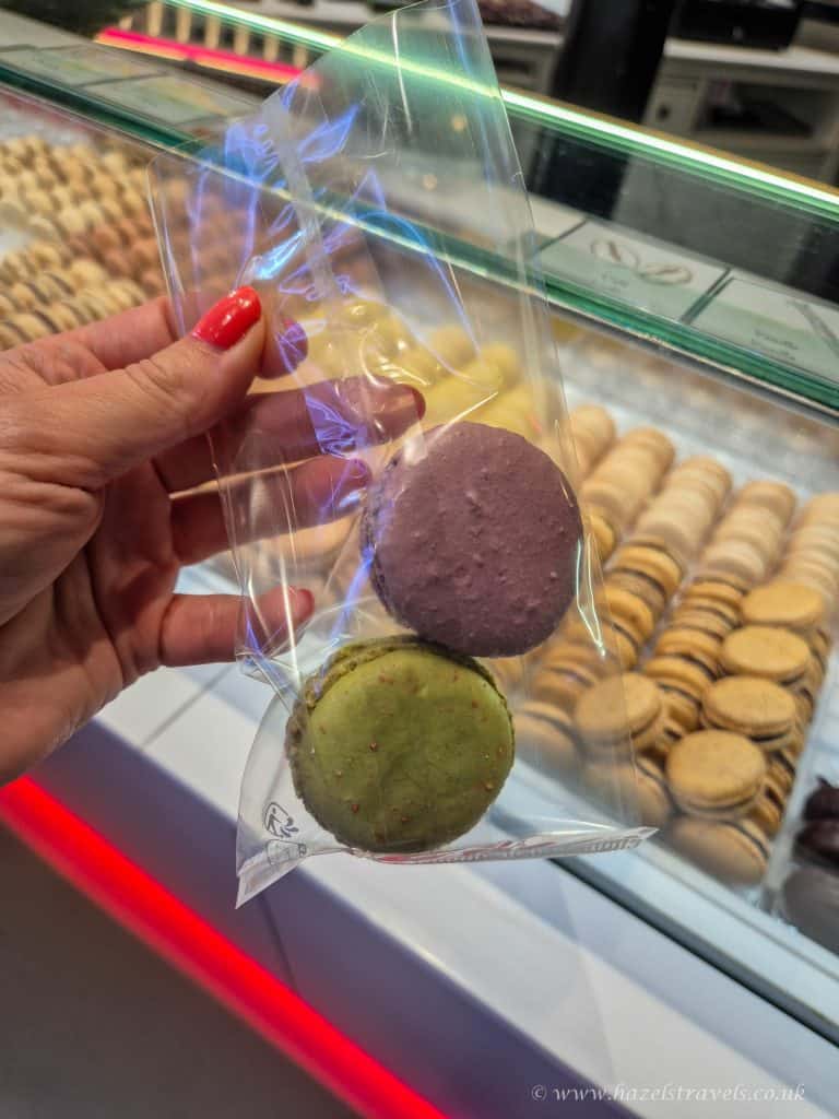 A hand holds a clear bag with two macarons, one purple and one green, in front of a display case filled with assorted macarons.