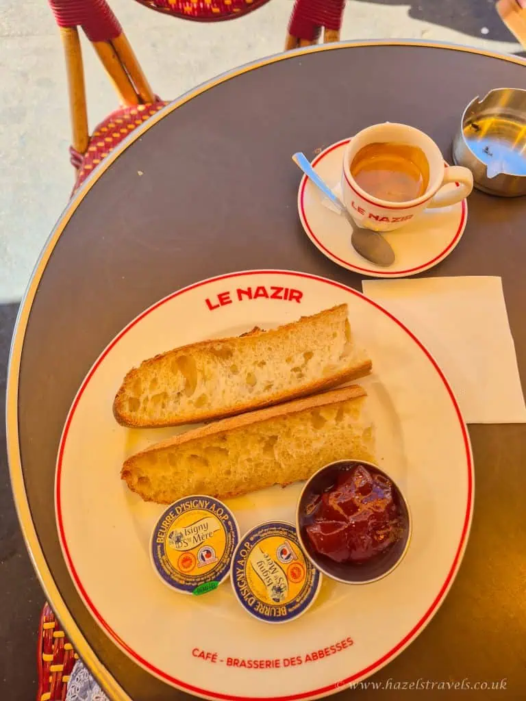 A table with a cup of espresso, a plate with two baguette slices, two butter portions, and a jam portion at Le Nazir café.