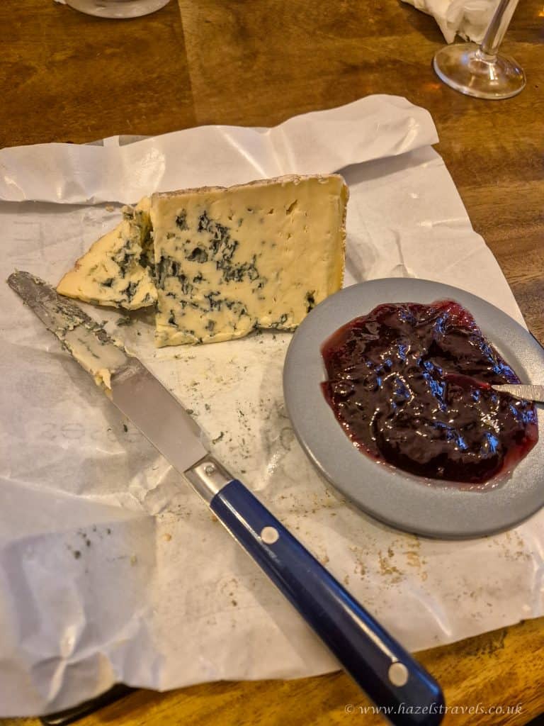 A wedge of blue cheese on parchment paper with a spreading knife and a small bowl of dark fruit jam on a wooden table.
