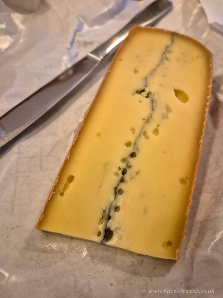 A wedge of Morbier cheese with a central line of ash, resting on parchment paper beside a metal knife.