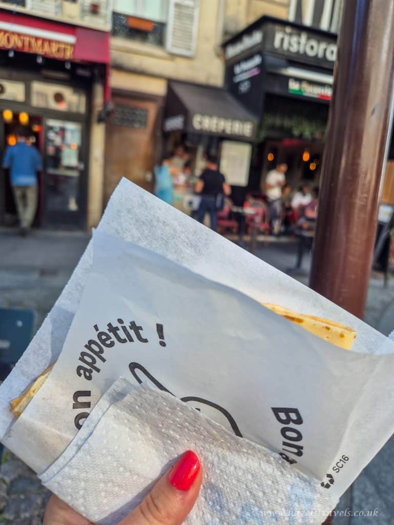 A hand with red nail polish holds a wrapped crepe in front of cafes and restaurants on a cobblestone street in a city.