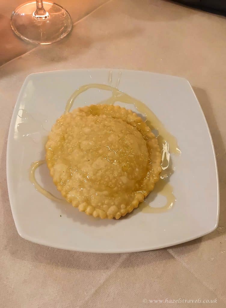 A single round fried pastry drizzled with honey sits on a square white plate atop a beige tablecloth, with part of a wine glass visible in the corner.