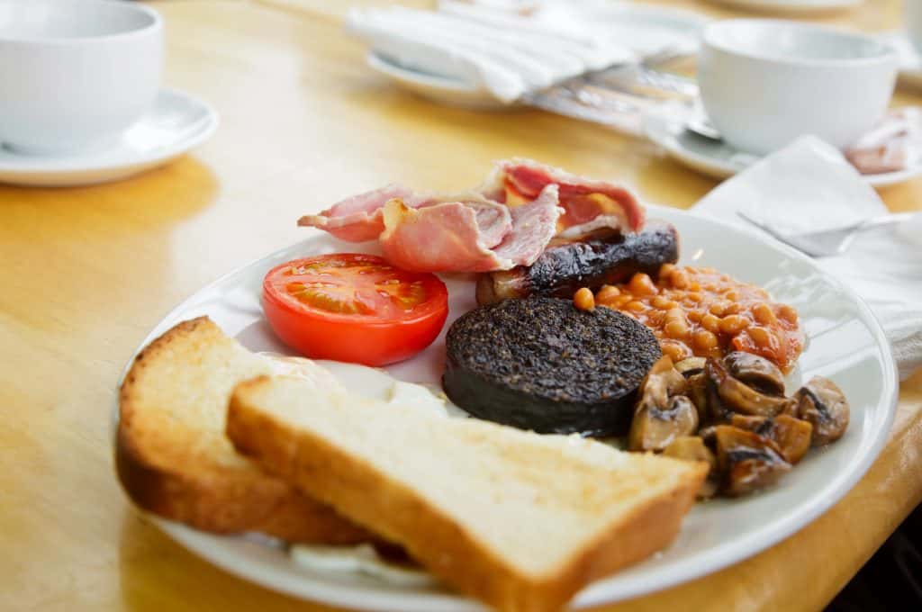 A full scottish breakdfast on a plate - toast, grilled tomato, bacon, black pudding, sausage, baked beans, and mushrooms on a wooden table, with cups and cutlery in the background.