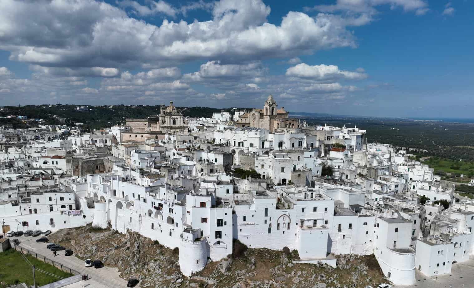 White hillside townscape of Ostuni, Puglia, Italy under a partly cloudy sky, featuring historic architecture, cobbled streets, and stunning views of the surrounding countryside.