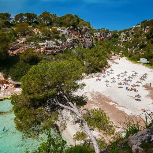 Turquoise beach with sun umbrellas in a secluded cove, surrounded by rocky cliffs and lush greenery.