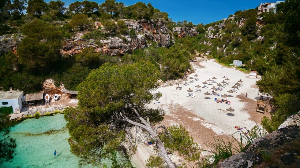 Turquoise beach with sun umbrellas in a secluded cove, surrounded by rocky cliffs and lush greenery.