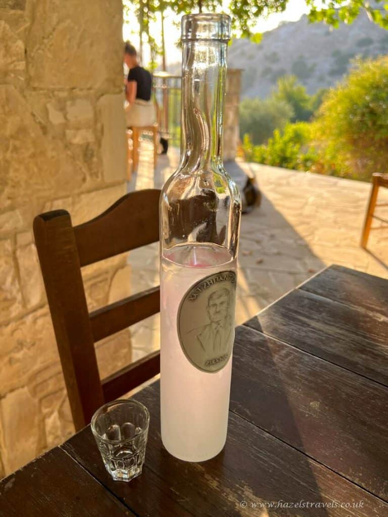 A tall frosted bottle with a silver label and a shot glass sit on a wooden table outdoors in sunny, rustic surroundings.