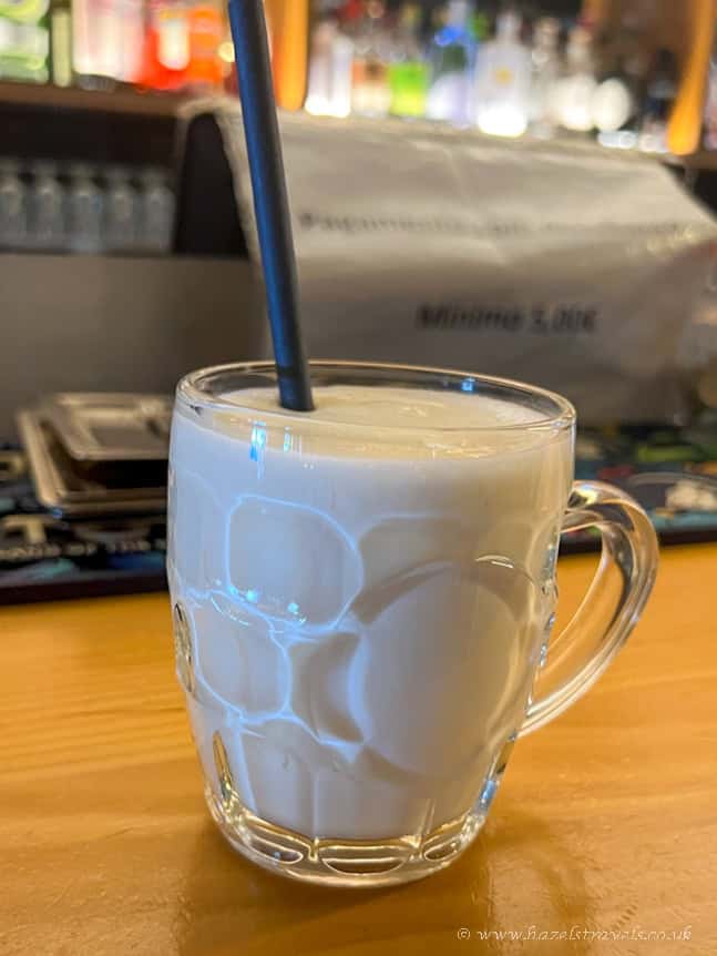 Hot chocolate in a glass mug with a straw on a wooden table, cosy café setting, warm drink, comfort, hazel's travels, UK travel, leisure, beverage, cafe scene, relaxing ambiance.