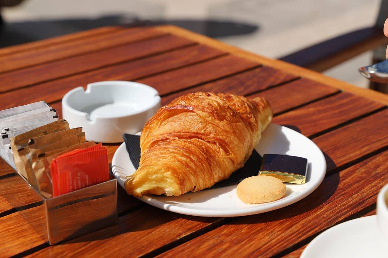 A freshly baked croissant with coffee and condiments on a wooden table.