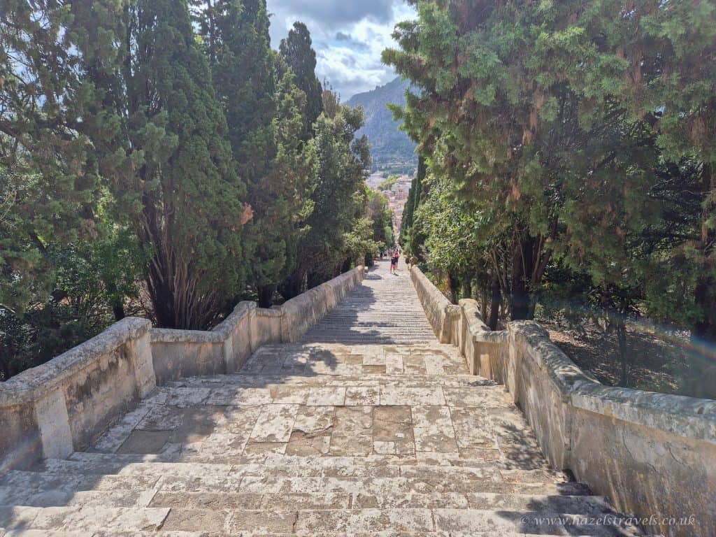 Stone staircase flanked by tall trees, descending towards a distant town with mountains in the background under a partly cloudy sky.