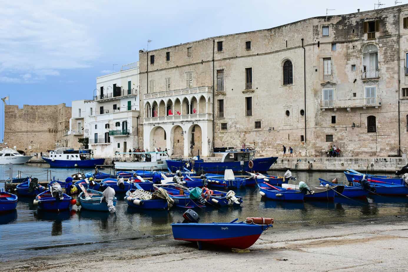 Colourful boats docked at a historic Mediterranean harbour with ancient stone buildings in the background.