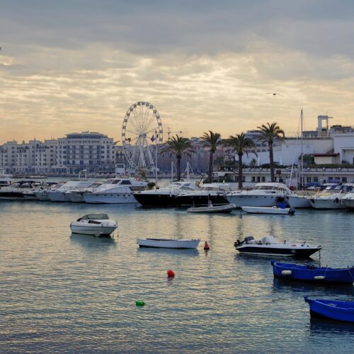 Boats docked at a harbour in a scenic lakeside city with a ferris wheel and modern architecture.