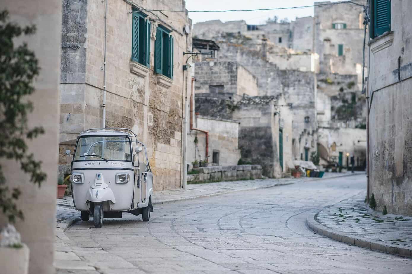 Vintage electric tuk-tuk parked on a cobbled street in an old Mediterranean village with stone buildings and green shutters, capturing charming travel moments in Europe.