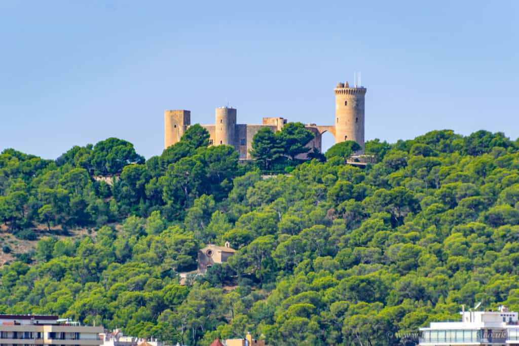 Medieval castle on green hilltop in Mallorca with lush trees and scenic landscape, under a clear blue sky.