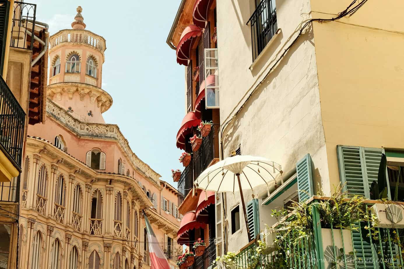 Colorful Mediterranean building facades with shutters, balconies, and a historic tower in the background, sunny day in an Italian or southern European city.