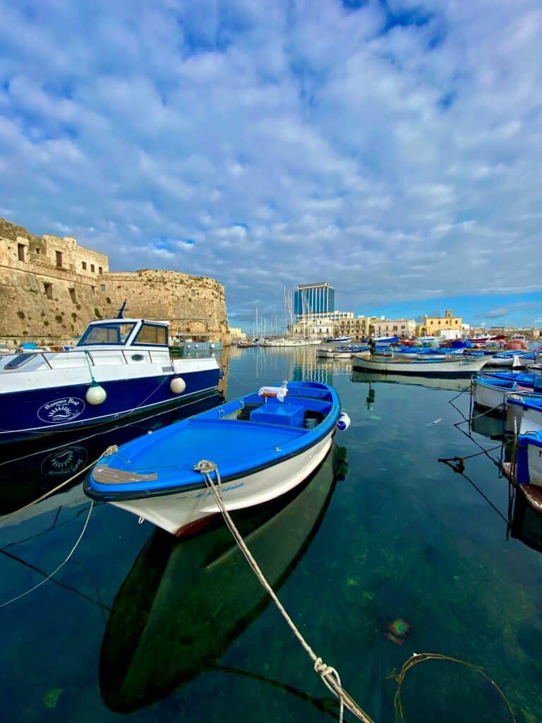 Colourful boats moored in a scenic marina with historic fortress walls and modern buildings in the background.