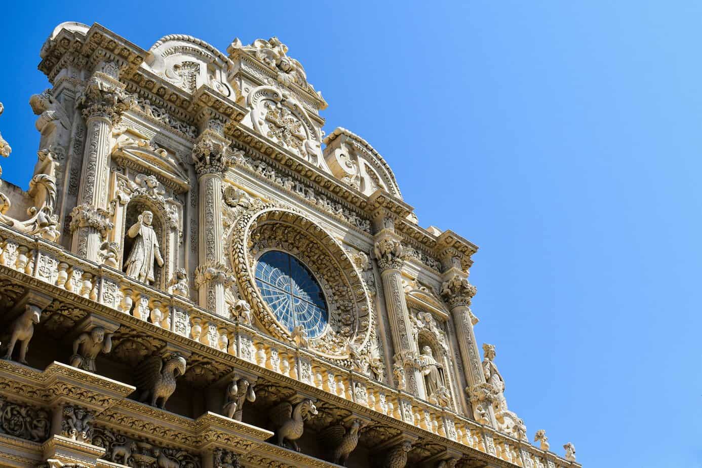 Ornate historic church facade with intricate stone carvings, sculptures, and a large circular stained glass window against a bright blue sky, highlighting European architecture and travel destinations.