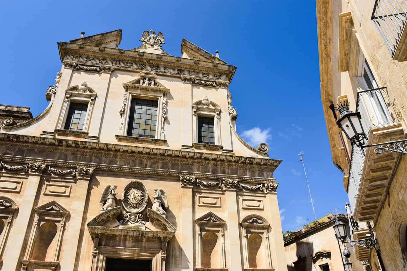 Ornate historic church facade with classical architecture in sunny Lecce in Puglia, featuring decorative stonework, angel sculptures, and elegant windows against a clear blue sky.