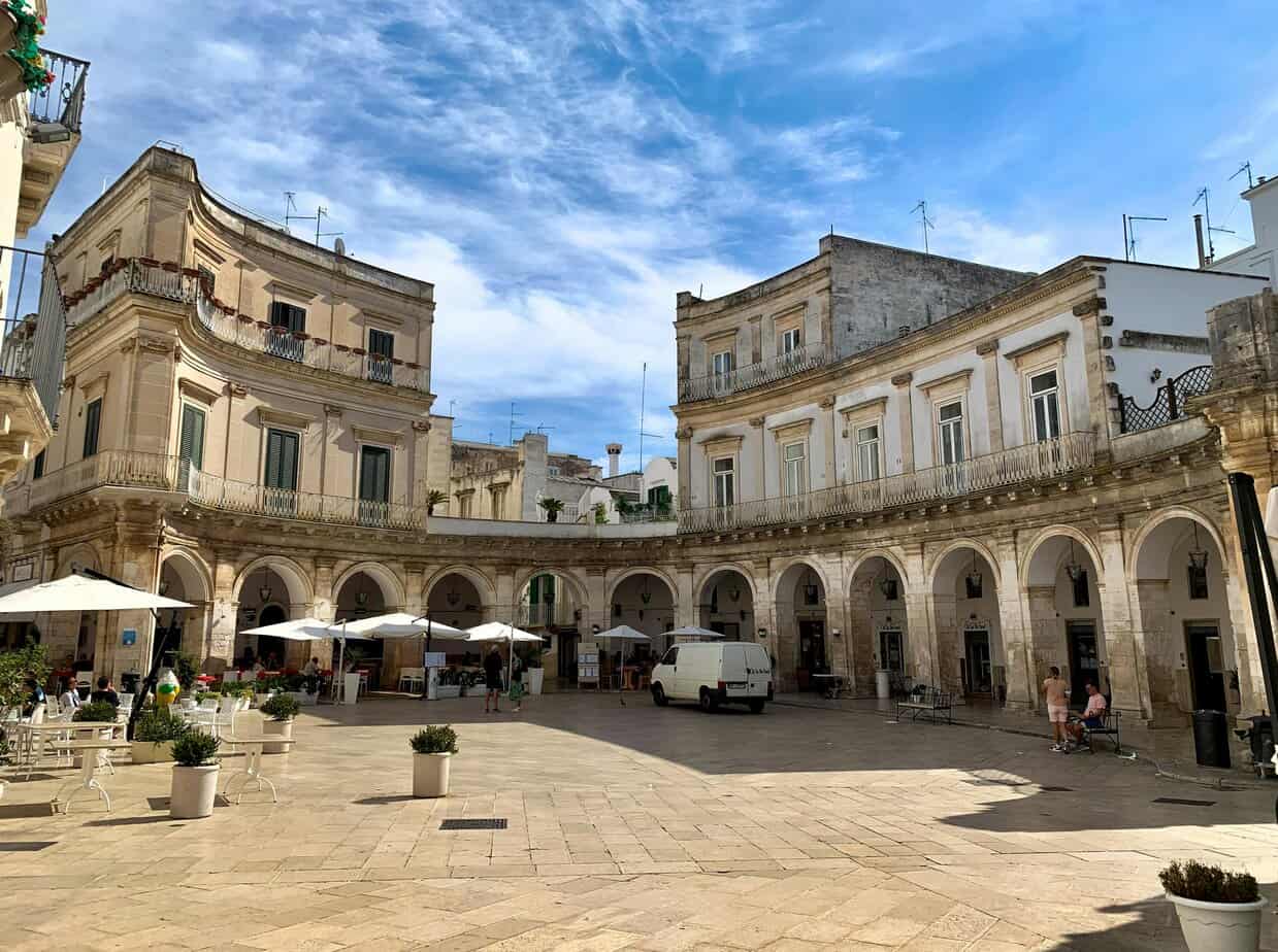 A historic Italian piazza with elegant white and beige buildings, open-air cafes with white umbrellas, and a bright blue sky.
