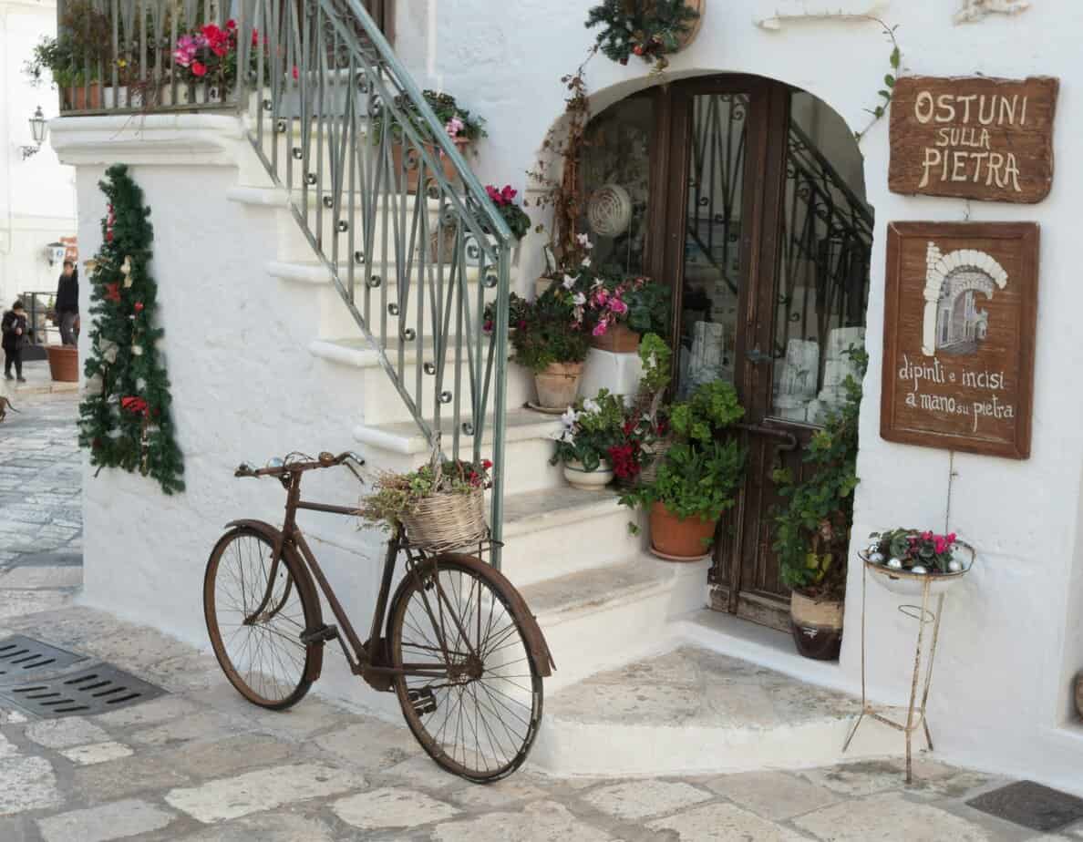 Vintage bicycle with flower basket outside white stone building in Italy's Ostuni, featuring decorative potted plants and traditional stone signage for handmade crafts.
