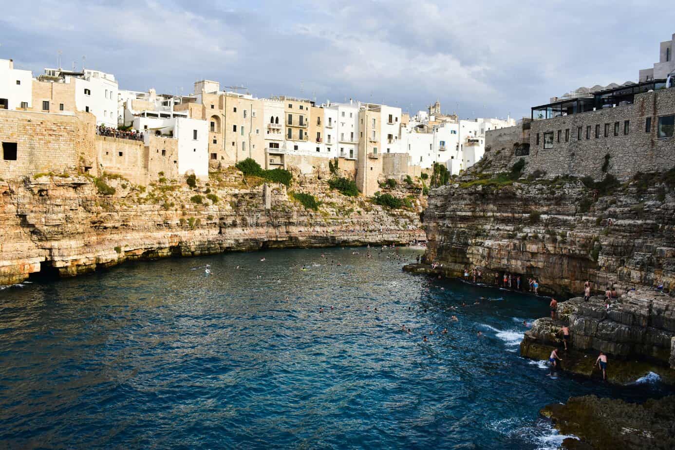 Seaside cliffside with white buildings overlooking a deep blue swimming cove in a Mediterranean coastal town.