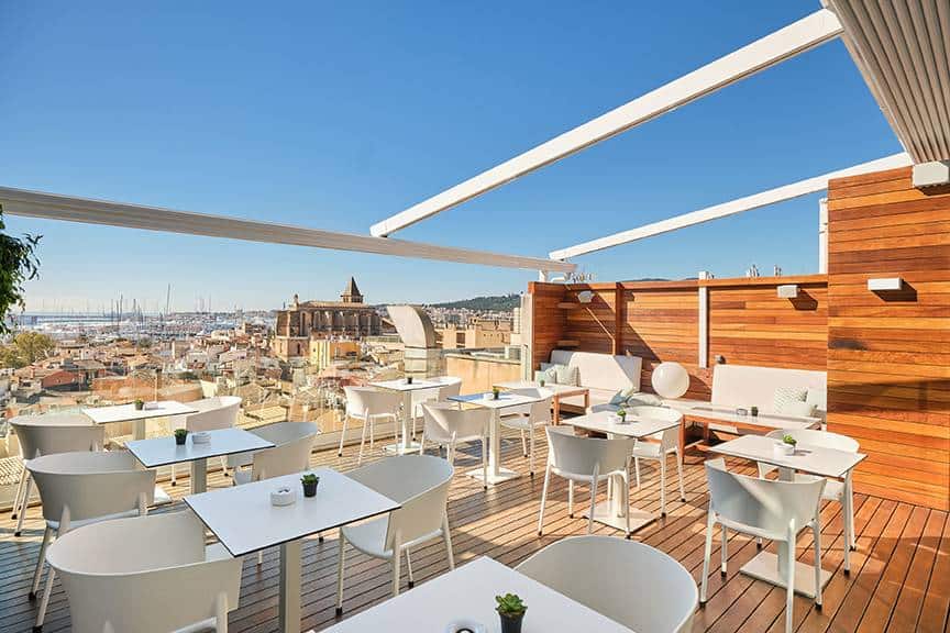 Rooftop terrace bar in Palma de Mallorca with modern white furniture, wooden decking, and panoramic views of the city, cathedral, and marina under a clear blue sky.