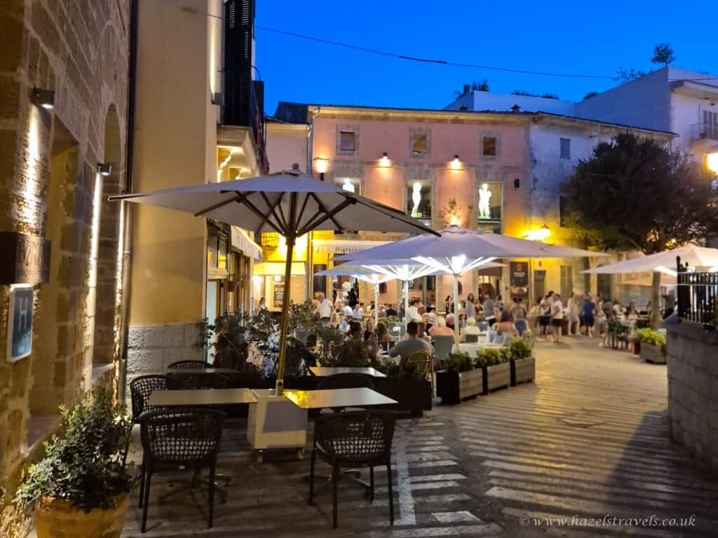 Evening scene in Alcudia Old Town, Mallorca, with people dining under white umbrellas at an outdoor restaurant surrounded by historic buildings and warm lights.