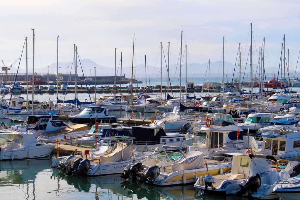 Scenic view of Alcudia Port in Mallorca with rows of sailboats and yachts moored in the marina, calm blue water, and distant mountains under a clear sky.