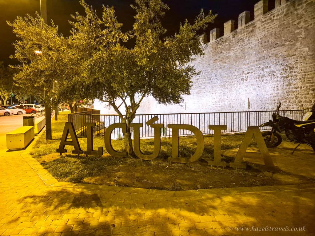 Night view of the illuminated Alcudia sign in front of the historic town walls and olive trees in Alcudia, Mallorca.