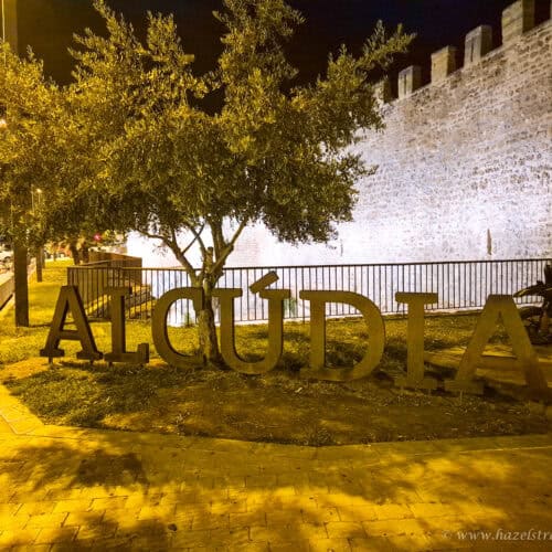 Night view of the illuminated Alcudia sign in front of the historic town walls and olive trees in Alcudia, Mallorca.
