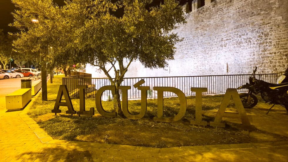 Night view of the illuminated Alcudia sign in front of the historic town walls and olive trees in Alcudia, Mallorca.