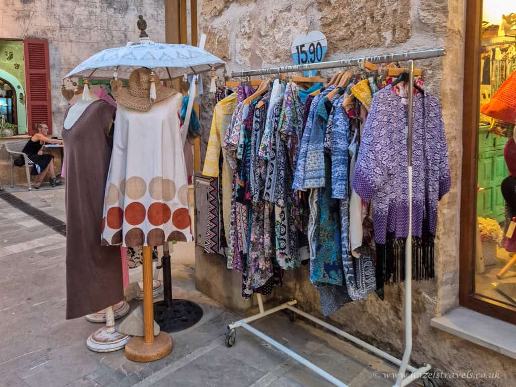 Colorful display of summer dresses and patterned clothing outside a boutique in Alcudia Old Town, Mallorca, set against a rustic stone wall.
