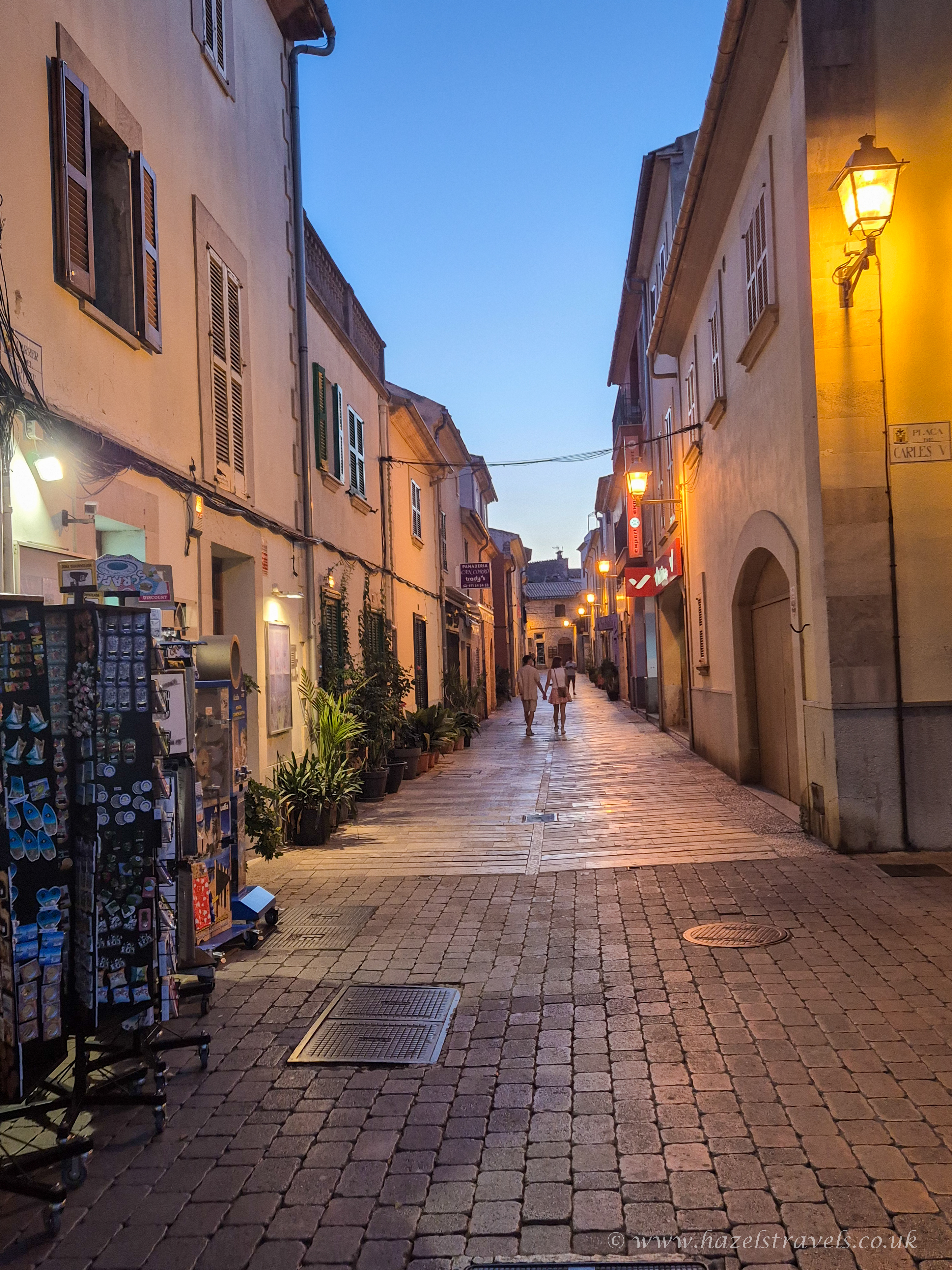 Narrow cobblestone street in Alcudia Old Town, Mallorca, at dusk with warm streetlights, historic buildings, and a few people strolling in the distance.