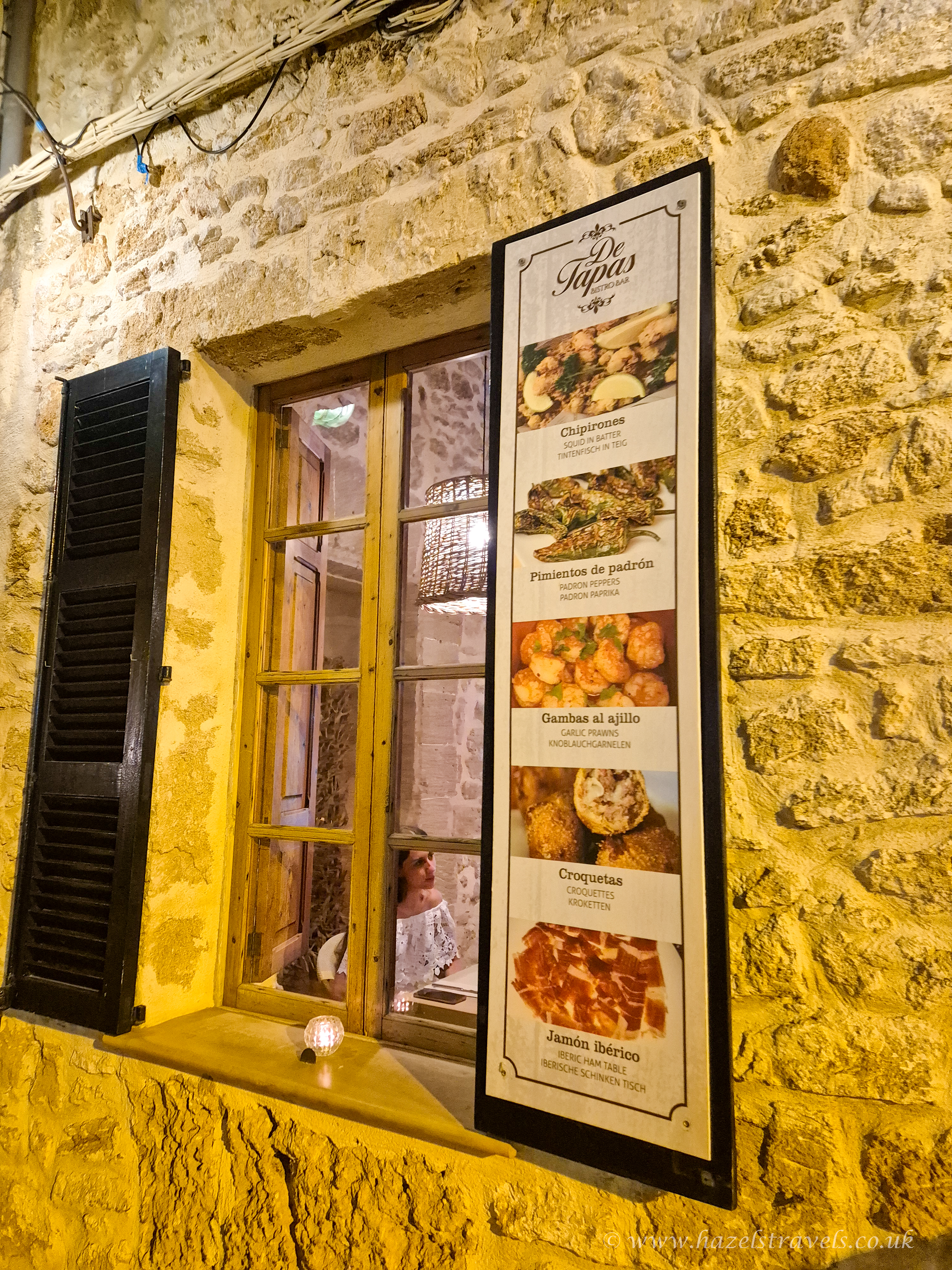 Rustic stone wall of a tapas bar in Alcudia, Mallorca, featuring a window with warm lighting and a vertical menu displaying traditional Spanish dishes like chipirones, padrón peppers, garlic prawns, croquetas, and jamón ibérico.