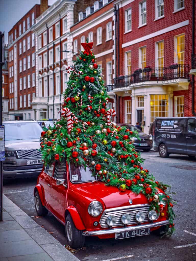 A classic red Mini car decorated with a giant Christmas tree and baubles parked on a festive London street.