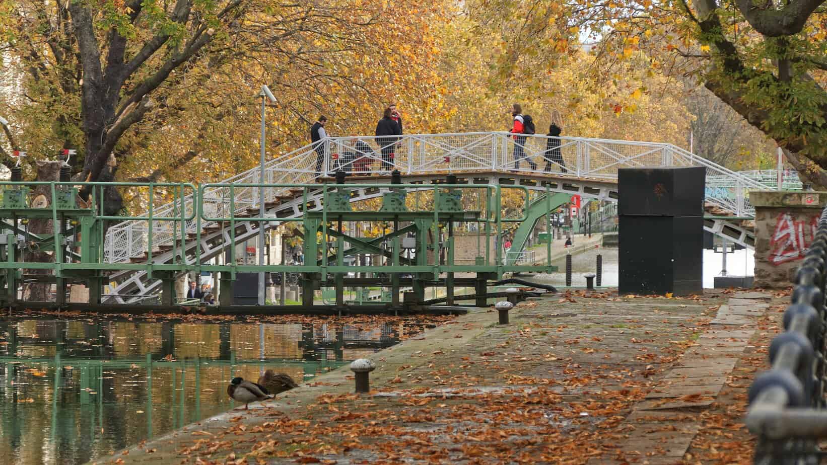 Footbridge over Canal Saint-Martin, Paris - People crossing the green iron bridge surrounded by autumn leaves.