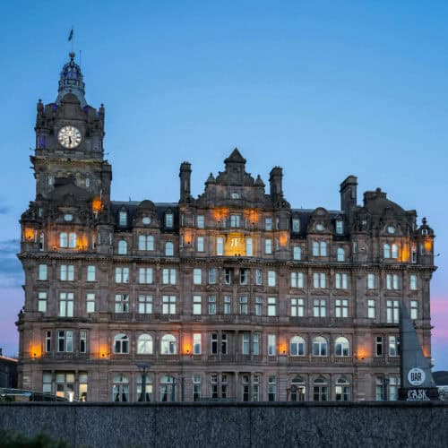 The Balmoral Hotel in Edinburgh illuminated at dusk, with its clock tower glowing against a clear blue sky.