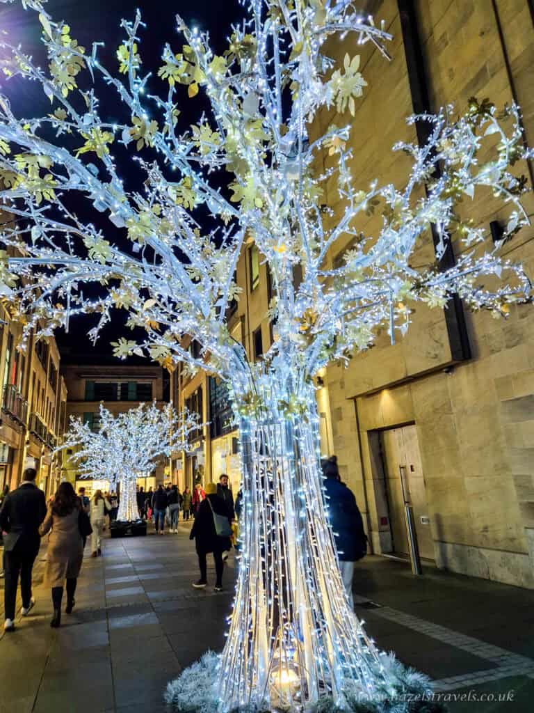 Illuminated tree covered in white and blue lights glowing along a cobbled Edinburgh street at night.