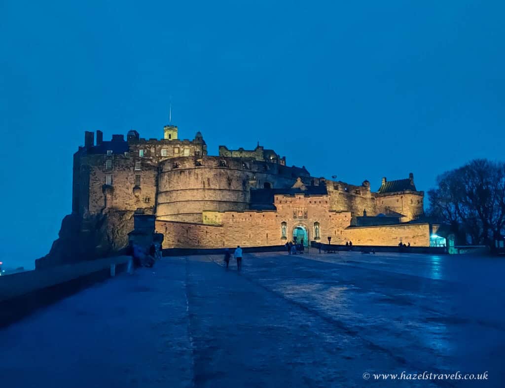 Edinburgh Castle illuminated at dusk, glowing golden against a deep blue evening sky.