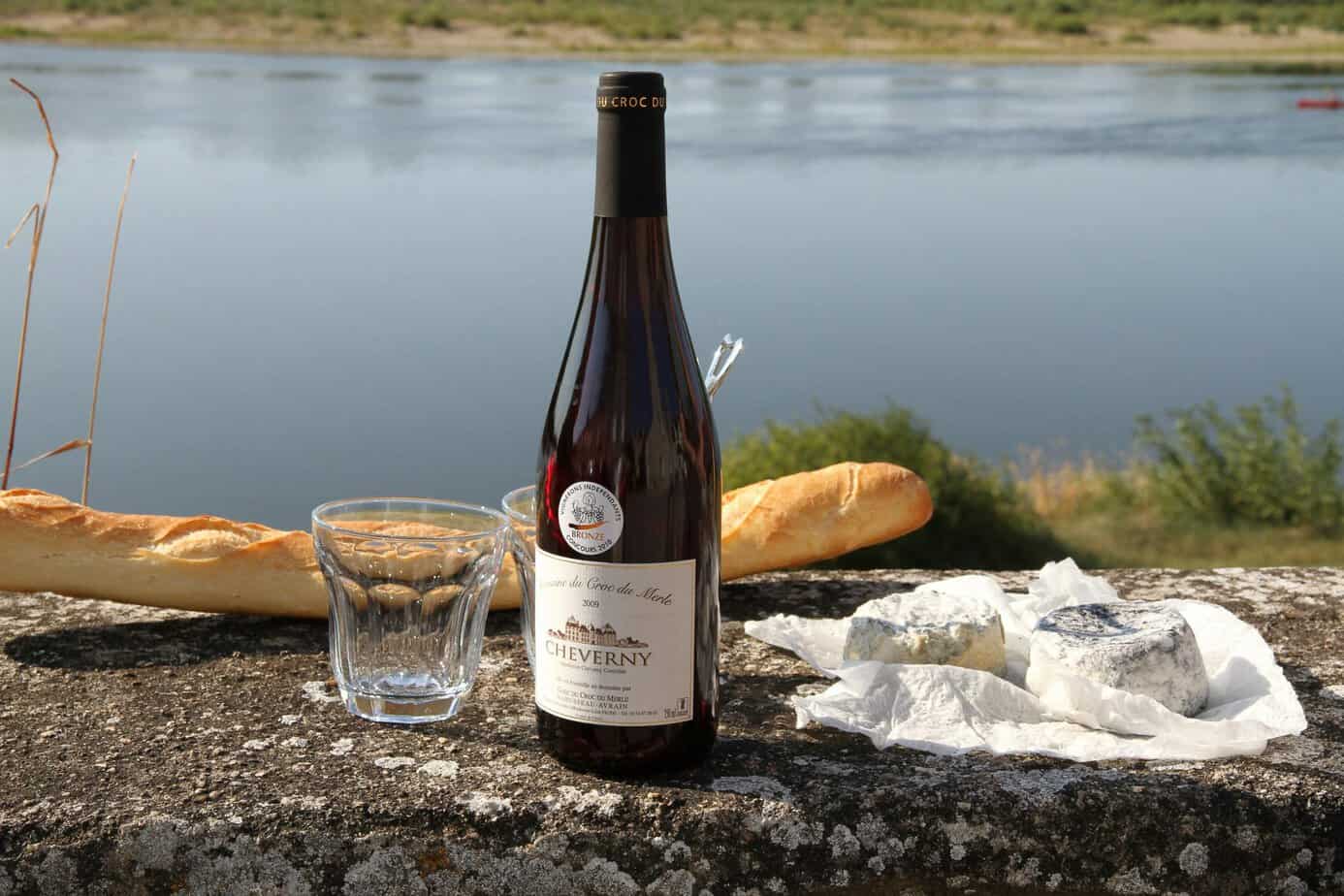 Wine and cheese by the river, France — Bottle of red wine, fresh cheese, and a baguette arranged on a stone ledge beside calm water.