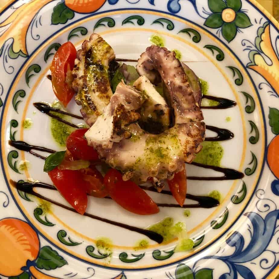 Fresh octopus tossed with parsley, lemon and olive oil, on a pattered plate with a balsamic drizzle and plum tomatoes.