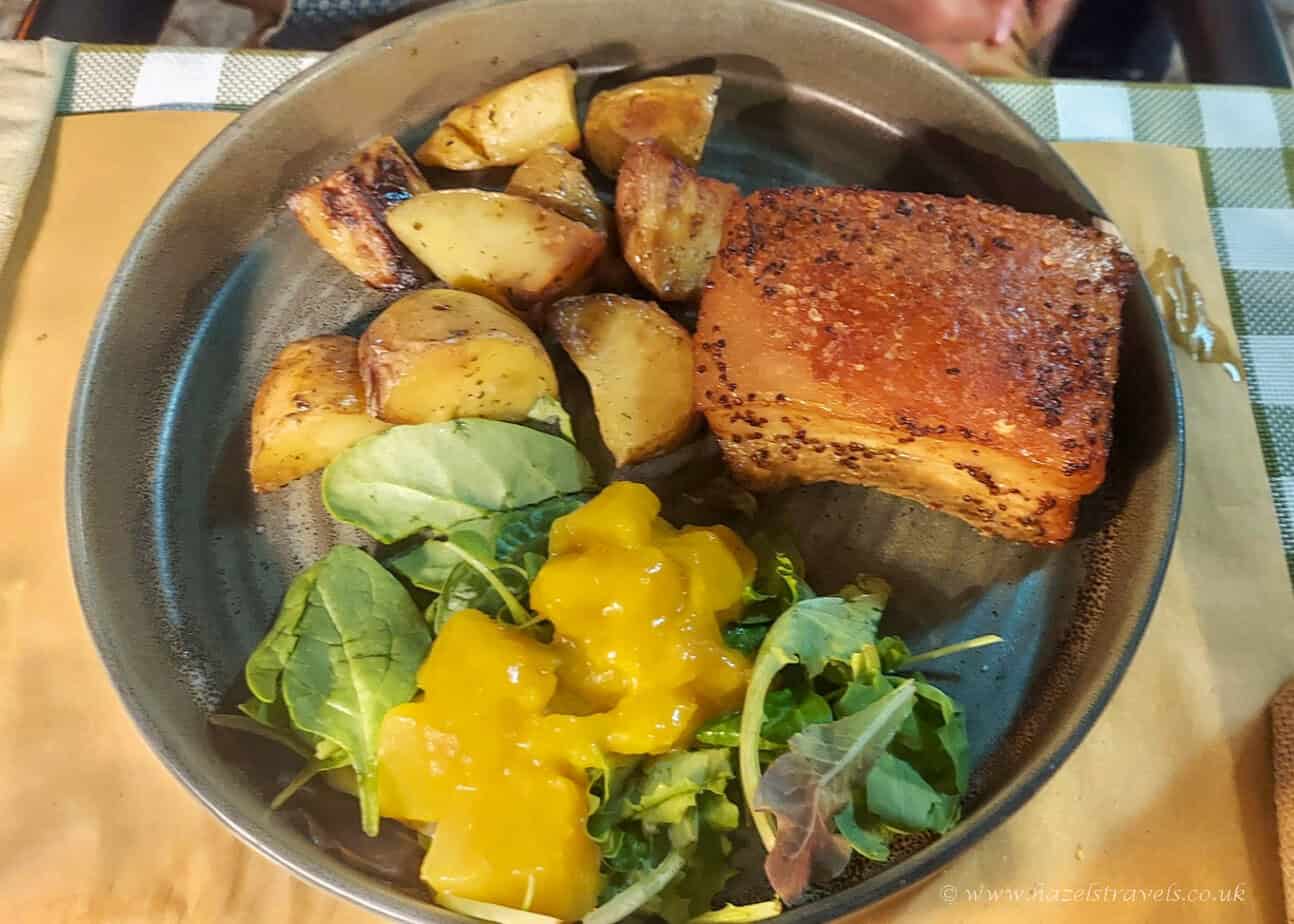 Plate of roasted pork belly with crispy crackling, served with golden potatoes, fresh salad leaves, and a dollop of mango chutney at a Sardinian restaurant.