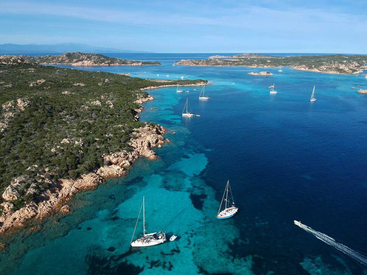 Aerial view of the La Maddalena Archipelago, Sardinia, showing turquoise bays, rocky coastlines, and sailing boats scattered across the clear blue sea.