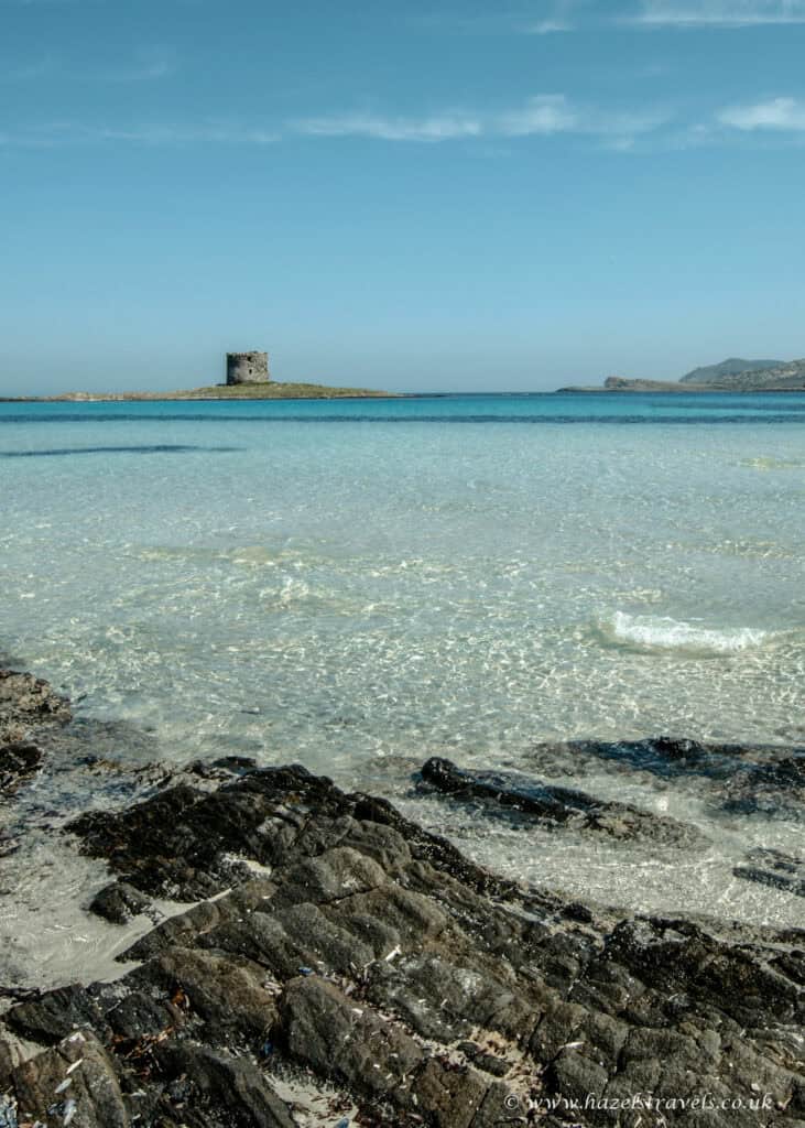 La Pelosa Beach, Sardinia - Shallow crystal-clear sea with Torre della Pelosa in the distance and rocky shoreline in the foreground.