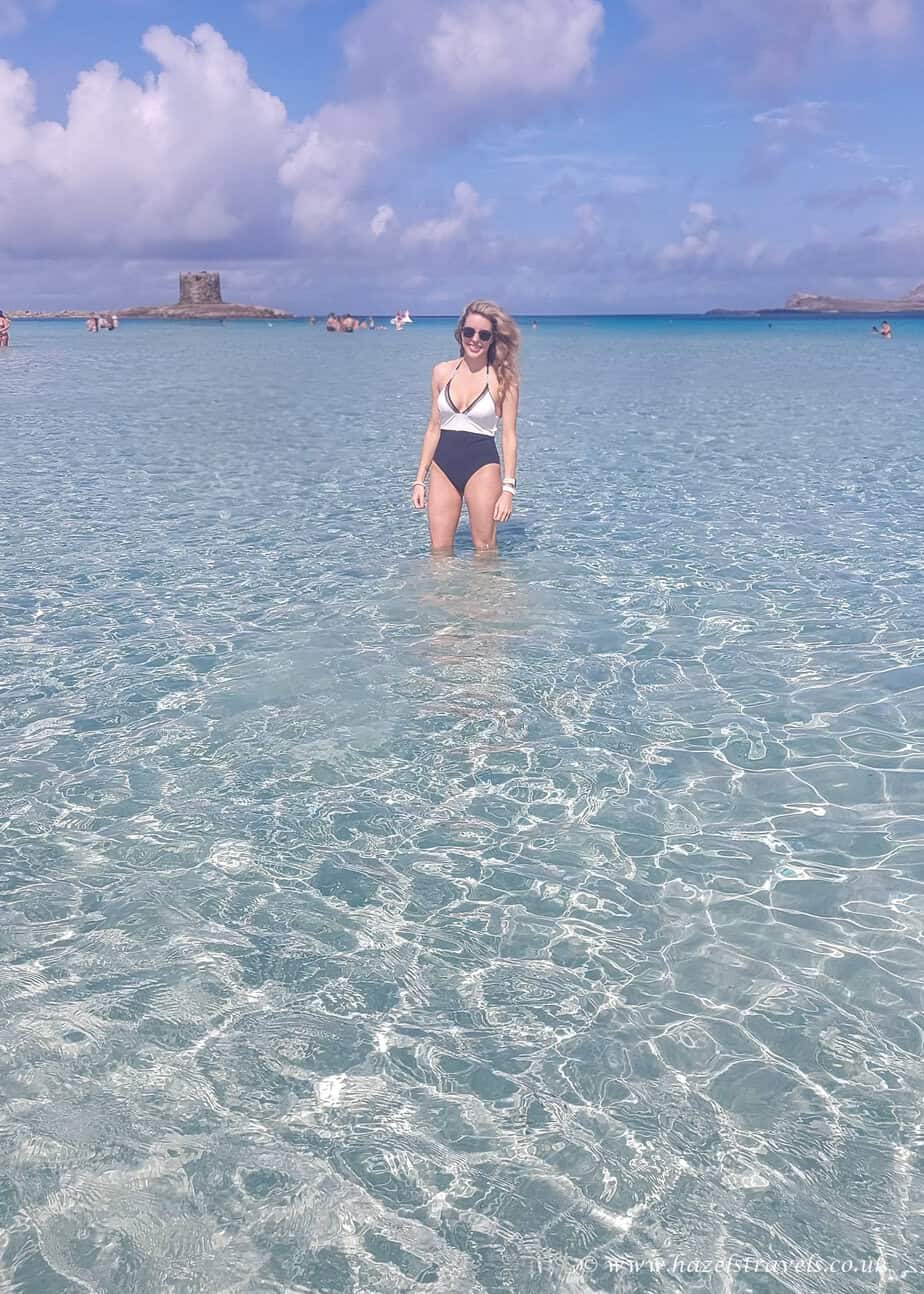 Wading in La Pelosa’s clear shallows, Sardinia - Woman standing in knee-deep, transparent water with the tower in the distance.